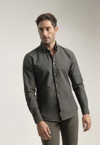 Camisa abotonada de color verde oscuro con cuello, mangas largas y una textura sutil. Presenta botones de color claro y un pequeño detalle de logo.
