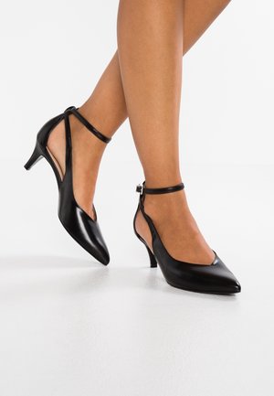 Ben iført sorte kitten heels med spids tå og ankelremme, stående på en glat hvid overflade.