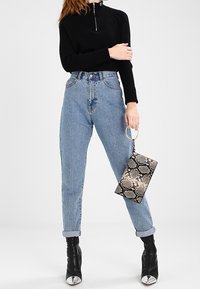 Pull à col zippé noir, jean taille haute bleu clair, bottines noires à bout argenté, tenant une pochette texturée en motif serpent.