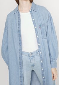 Vestido de camisa de denim azul claro con mangas largas abullonadas, cierre de botones en el frente y un bolsillo en el pecho. Combinado con una blusa blanca y jeans.