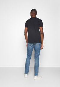 Camiseta negra de manga corta, fabricada en algodón, combinada con vaqueros ajustados azul claro. Modelo de espaldas, lleva zapatillas blancas, fondo neutro.