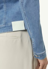 Dettaglio di una manica di giacca di denim azzurro chiaro con un'etichetta bianca strutturata sul retro, accanto a un indumento color crema.