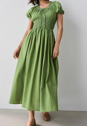 Kobieta w zielonej sukience midi z bufkami na rękawach, marszczonym gorsetem i zwiewną spódnicą, w zestawieniu z brązowymi, plecionymi butami wsuwanymi.