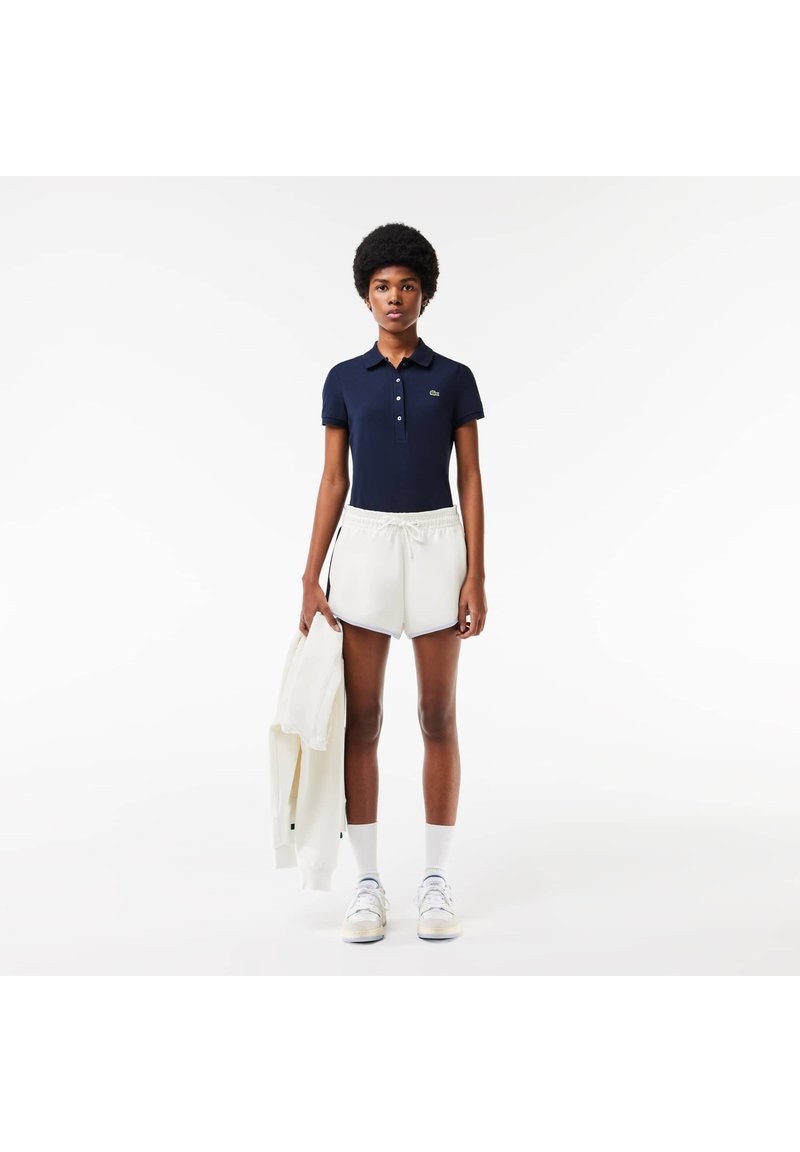 Marineblauwe polo met korte mouwen en een kraag, gecombineerd met witte korte broek met trekkoord. Witte sweatshirt over de linkerarm gehangen. Witte sneakers.