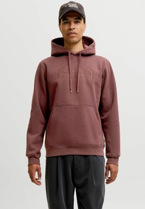 Mężczyzna ubrany w brązową bluzę z kapturem Jack & Jones Premium i czarną czapkę Jack & Jones stoi na jednolitym jasnym tle.