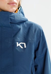 Blaue Outdoorjacke mit hohem Kragen, mit weißem Logo auf der linken Seite und glatter Textur. Kapuze inklusive, funktional gestaltet.
