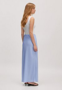 Lichtblauwe satijnen maxi-rok met een gladde textuur, gecombineerd met een aansluitend wit tanktop. Model wordt van achteren weergegeven, met zwarte slippers.