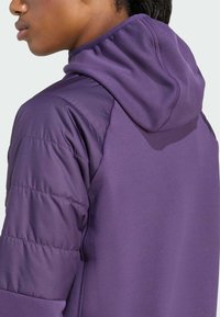 En närbild av en persons axel och nacke, iklädd en lila huva jacka. Tyget visar en blandning av släta och vadderade texturer.