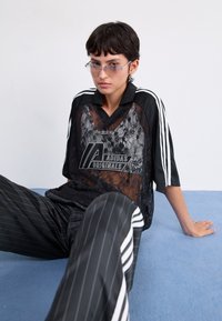Czarna koronkowa koszulka Adidas Originals z czarnym kołnierzykiem, z logo i paskowanymi spodniami w czarno-białe prążki. Noszone okulary przeciwsłoneczne.