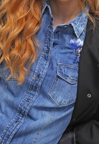 Lang rood haar over een blauw denim shirt met witte en blauwe bloemen borduurwerk op de borstzak, gelaagd onder een zwarte jas.