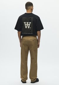 T-shirt noir surdimensionné avec un grand logo blanc au dos, associé à un pantalon marron, porté avec des baskets noires. Tissu lisse, coupe décontractée.