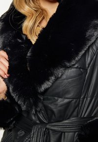 Czarna pikowana kurtka z miękkim czarnym kołnierzem z futra ekologicznego, paskiem i prążkowanymi detalami na tkaninie o gładkiej fakturze.