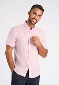 Camicia a maniche corte rosa con bottoni realizzata in tessuto testurizzato, caratterizzata da un colletto e un orlo curvo. Indossata in modo informale con pantaloni scuri.
