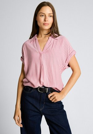 Kobieta w różowej bluzce w paski z krótkim rękawem, włożonej do ciemnoniebieskich dżinsów z czarnym paskiem, stojąca na jednolitym tle.