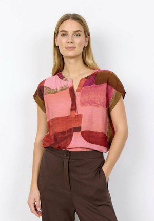 Vrouw draagt een blouse met korte mouwen, multicolor roze en bruin abstract patroon, en donkerbruine, hooggetailleerde broek, hand in de zak.