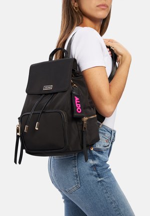 Black nylon backpack with gold hardware, adjustable straps, front pocket, and a pink "ALDO" tag. Features a fold-over flap with drawstring closure.