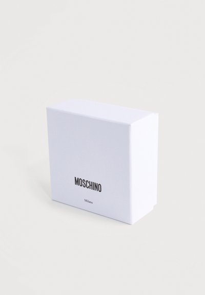 Boîte rectangulaire blanche avec texte noir "MOSCHINO" et texte plus petit "Milano" à l'avant, placée sur un fond neutre clair.