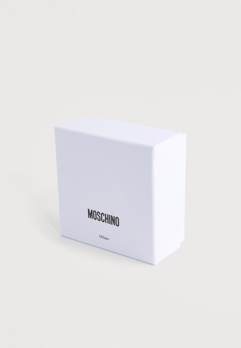 Boîte rectangulaire blanche avec texte noir "MOSCHINO" et texte plus petit "Milano" à l'avant, placée sur un fond neutre clair.