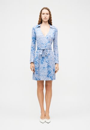 Kobieta stojąca w niebieskiej sukience kopertowej z białymi wzorami liści, z długimi rękawami i paskiem w talii, w połączeniu z białymi butami na obcasie z ostrym noskiem.