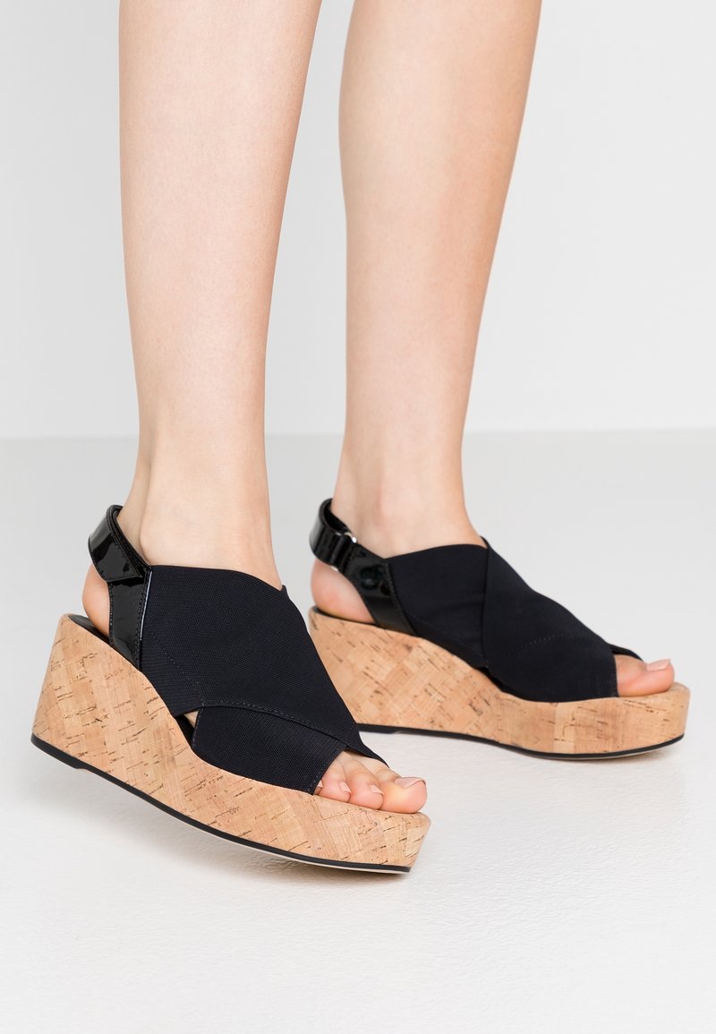 Sandales compensées noires avec dessus élastique et plateforme en liège. Elles présentent un design à bout ouvert et une sangle de cheville réglable avec des accents noirs brillants.