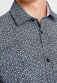 Marinblå skjorta med knappar som har ett litet vitt blommönster, spetsig krage och textil med struktur. Synlig knapplist och kontrasterande innerekrage.