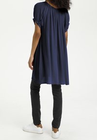 Tunique bleu marine avec un détail plissé au dos, manches courtes retroussées, associée à un pantalon noir et des baskets blanches, avec une coupe décontractée.