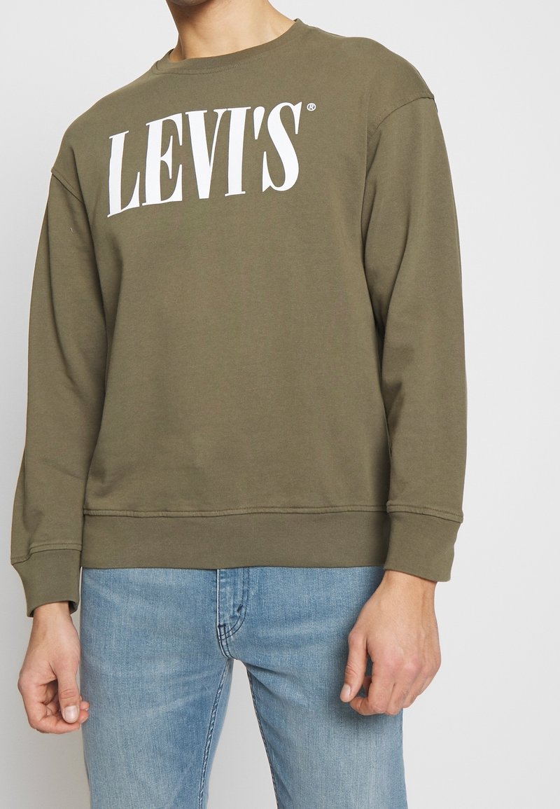 Homme portant un sweat-shirt vert olive Levi's et un jean bleu clair, debout devant un fond clair uni.