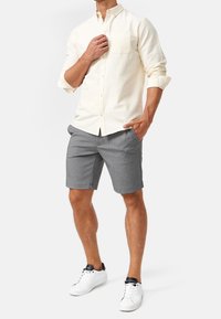 Lichtbeige lange mouw blouse met knoopsluiting en een borstzak, gecombineerd met grijze shorts en witte sneakers, staande tegen een grijze achtergrond.