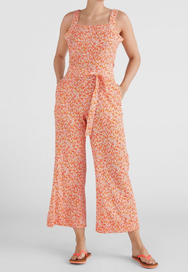 Femme portant une combinaison sans manches à imprimé floral avec des jambes larges, une ceinture assortie et des tongs corail, debout les mains dans les poches.