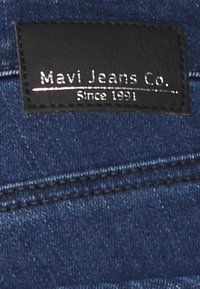 Etiqueta de couro preta com texto branco a dizer "Mavi Jeans Co." e "Desde 1991," cosida em tecido de ganga azul-escura.
