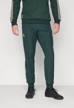 Grøn sports tracksuit med hvide sidestreger, med et logo på venstre ben. Stoffet ser blødt ud med en tætsiddende pasform. Sorte sko.