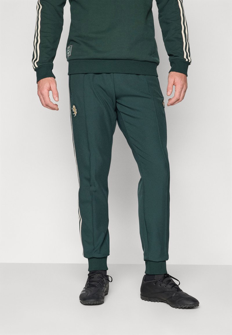 Grüner Sportanzug mit weißen Seitenstreifen, mit einem Logo auf dem linken Bein. Der Stoff wirkt weich mit einem körperbetonten Design. Schwarzfarbige Schuhe.