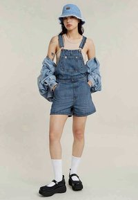 Woman wearing blue bucket hat, denim short overalls, beige tank top, oversized denim jacket, white socks, and black platform shoes.