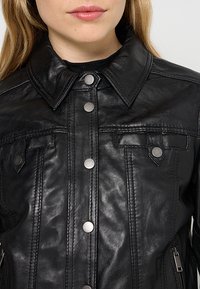 Veste en cuir noir avec boutons-pression, un col pointu et des poches frontales. Texture lisse avec des détails de couture visibles et une fermeture éclair asymétrique.