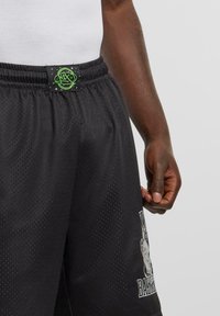 I pantaloni da basket neri in rete presentano una superficie testurizzata e una cintura elastica con un'etichetta con logo verde. Una stampa grafica decora il lato.