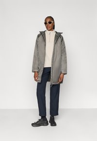 Cappotto puffy grigio lucido di media lunghezza con cappuccio, indossato sopra un maglione chiaro e pantaloni scuri; persona che indossa occhiali da sole oversize e sneakers.