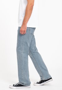 Jean en denim bleu clair avec une coupe droite, dotés de poches arrière et d'une finition douce et texturée. Porté avec des baskets noires et blanches.