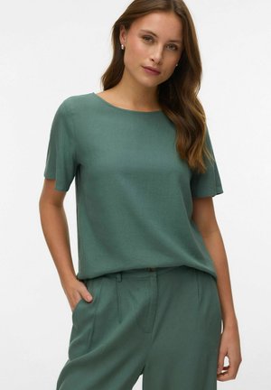 Kobieta o długich brązowych włosach, ubrana w zielony krótki top z krótkim rękawem i dopasowane zielone spodnie z wysokim stanem, stojąca z jedną ręką w kieszeni.