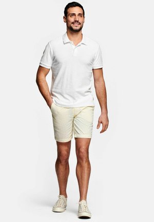 Mannelijk model lopend, gekleed in een wit poloshirt met korte mouwen, lichtbeige korte broek en witte sneakers tegen een effen lichte achtergrond.