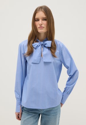 Mujer joven con blusa azul a rayas y un gran lazo en el cuello, y jeans azul claro, de pie con una mano en el bolsillo.