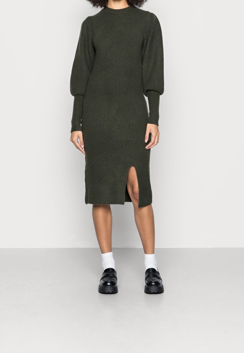 Robe tricotée vert olive avec texture côtelée, manches longues bouffantes, encolure ronde et fente latérale mi-cuisse. Paire de chaussures noires et chaussettes blanches.