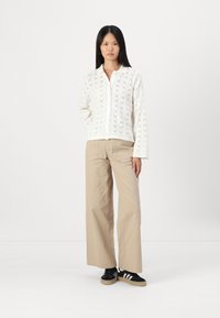 Bela pletena kardigan s cvetličnim vzorcem, bež široke hlače in črne superge z belimi črtami. Model stoji pred enobarvno ozadjem.