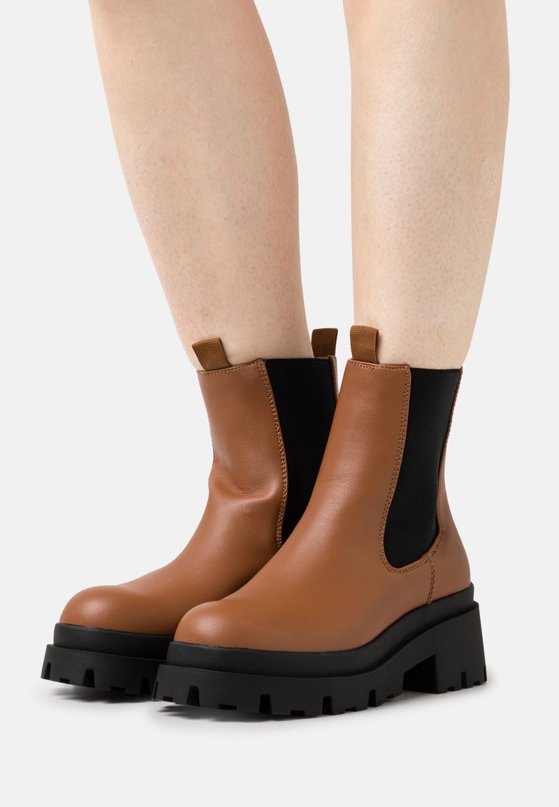 Bottes chevilles en cuir marron avec panneaux élastiques noirs sur les côtés et semelles épaisses noires portées sur des jambes nues sur un fond clair.
