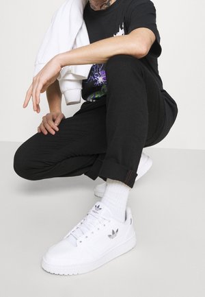 Persoon die hurkt, gekleed in een zwarte broek, een zwart T-shirt met een grafisch ontwerp, witte sokken, witte Adidas sneakers, en een witte jas over één schouder dragend.