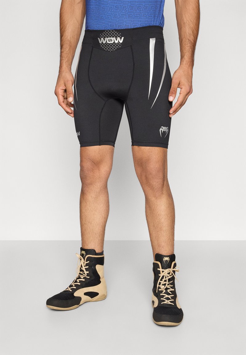 Calções de compressão pretos com detalhes prateados, com ajuste justo e cintura elástica. Usados com sapatilhas desportivas de cano alto em preto e castanho.