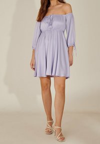 Vestido de lilas sin hombros con mangas de tres cuartos, cintura ajustada y falda fluida. Tejido suave con caída ligera. Un par de tacones beige.