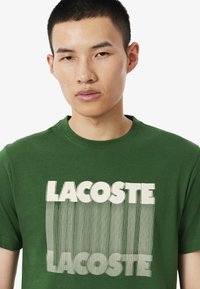 Camiseta de algodón verde con gran texto blanco "LACOSTE" y patrones de líneas verticales. Cuello redondo y mangas cortas, ajuste casual.