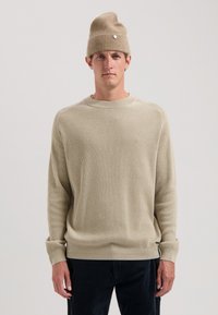 Beige geripptes Strickpullover mit rundem Ausschnitt und langen Ärmeln, kombiniert mit einer passenden beige Mütze mit einem kleinen metallischen Akzent.