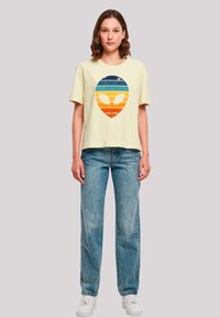 Yellow cotton t-shirt with a graphic alien design in blue and orange stripes. Paired with light blue denim jeans and white sneakers.