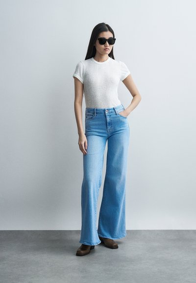 Γυναίκα με μαύρα γυαλιά ηλίου, άσπρο κοντομάνικο τοπ με υφή, ψηλόμεσα ανοιχτό μπλε τζιν παντελόνια φαρδιάς γραμμής και καφέ μποτάκια με τακούνι, στέκεται με το χέρι στην τσέπη.